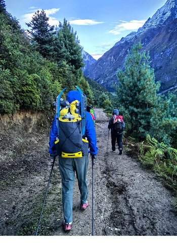 Two hikers with backpacks on a mountain trail with trees and mountains in the background.